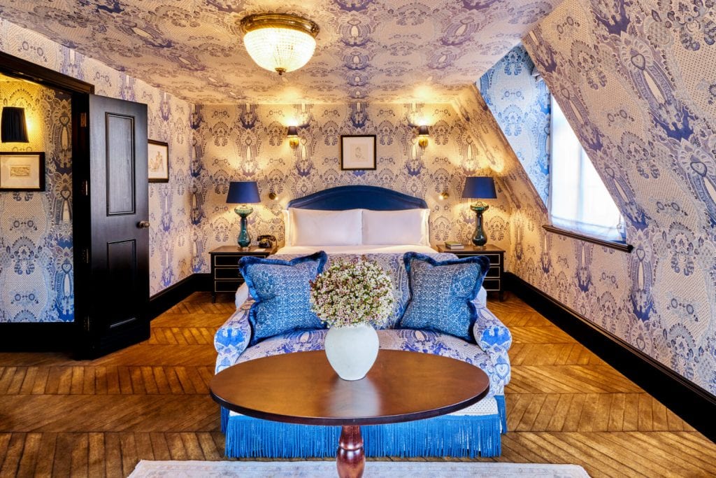 A cozy bedroom with blue patterned wallpaper, a double bed with a blue headboard, matching lamps and pillows, a floral sofa, a round table with a vase of flowers, wood flooring, and sloped ceiling.