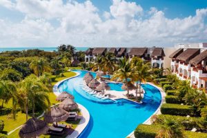 A tropical resort features a winding outdoor pool surrounded by palm trees, thatched cabanas, and sun loungers, with white buildings in the background under a bright blue sky with scattered clouds.