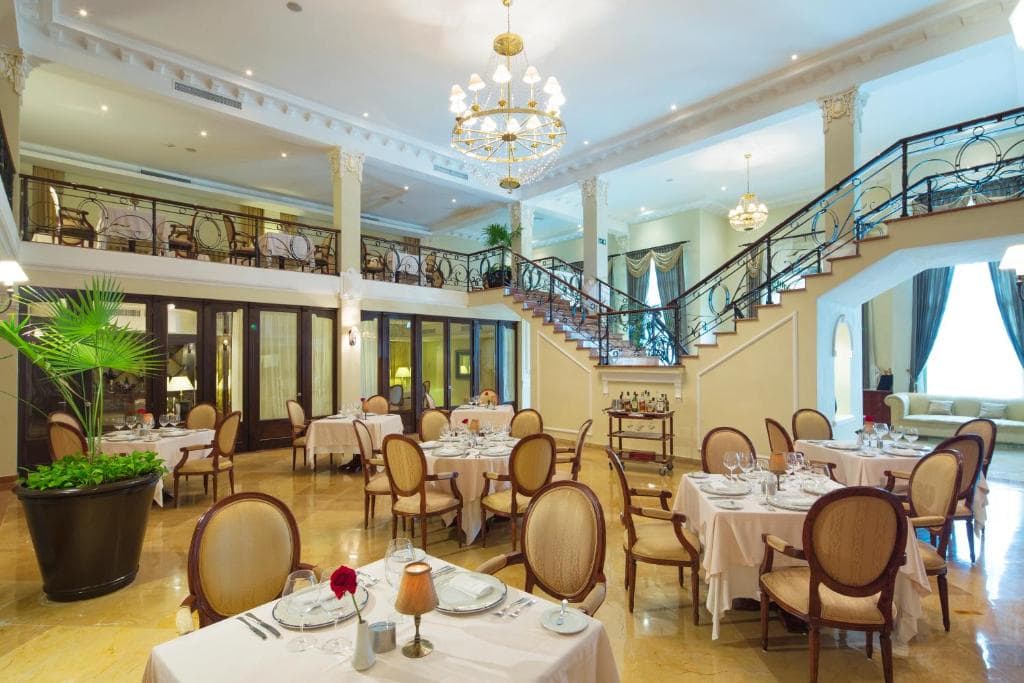 Elegant restaurant interior with round tables set for dining, upholstered chairs, chandeliers, large potted plants, marble floors, a sweeping staircase, mezzanine level, and natural light from tall windows with drapes.