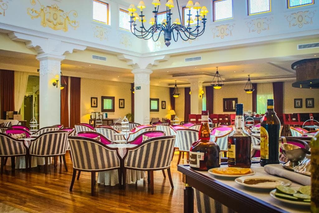 Elegant restaurant interior with striped chairs, round tables set with white tablecloths, and a chandelier hanging from a high ceiling. Bottles and plates are on a table in the foreground. Warm and inviting atmosphere.