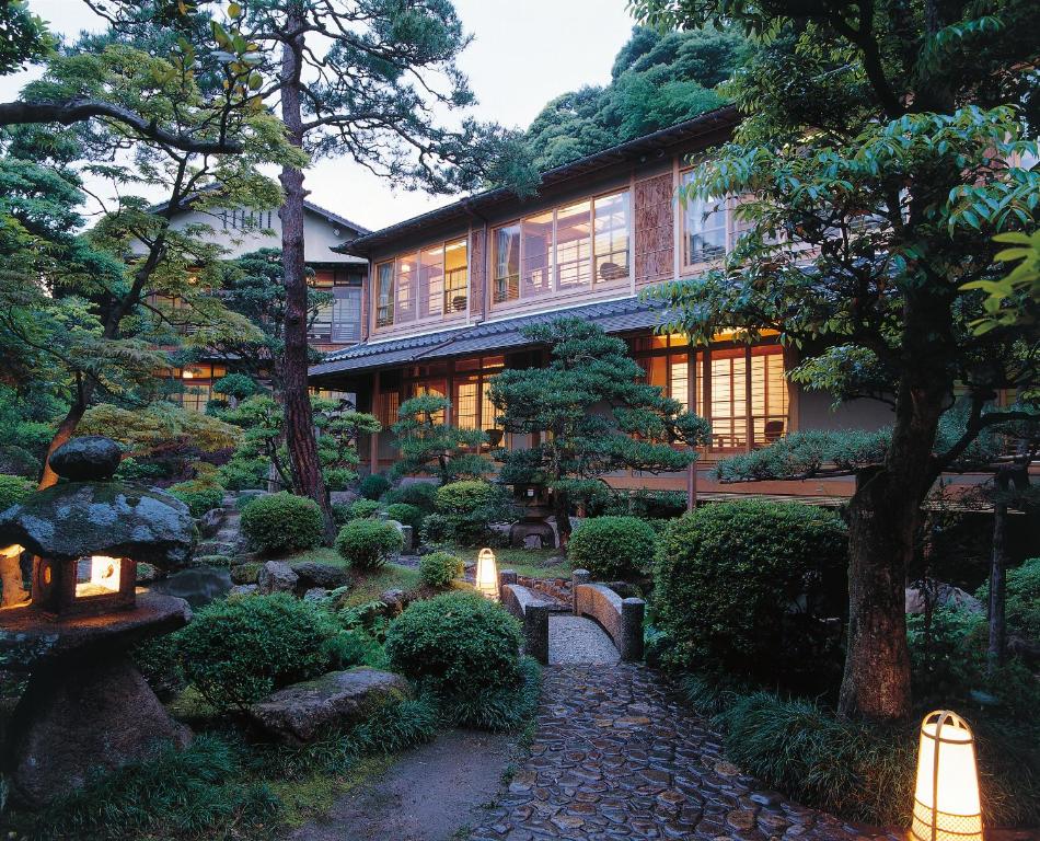 A traditional Japanese house with sliding doors and warm lights is surrounded by a lush garden, stone lanterns, and neatly trimmed bushes along a stone pathway at dusk.