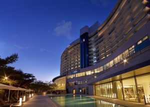 A modern, curved hotel with many balconies is lit up at dusk, overlooking a calm outdoor pool. Trees line one side, and lounge chairs with umbrellas are set up beside the water.
