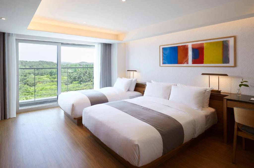 A modern hotel room with two neatly made beds, a large window overlooking greenery, a wooden floor, a desk with a chair, and a colorful abstract painting on the wall above the beds.