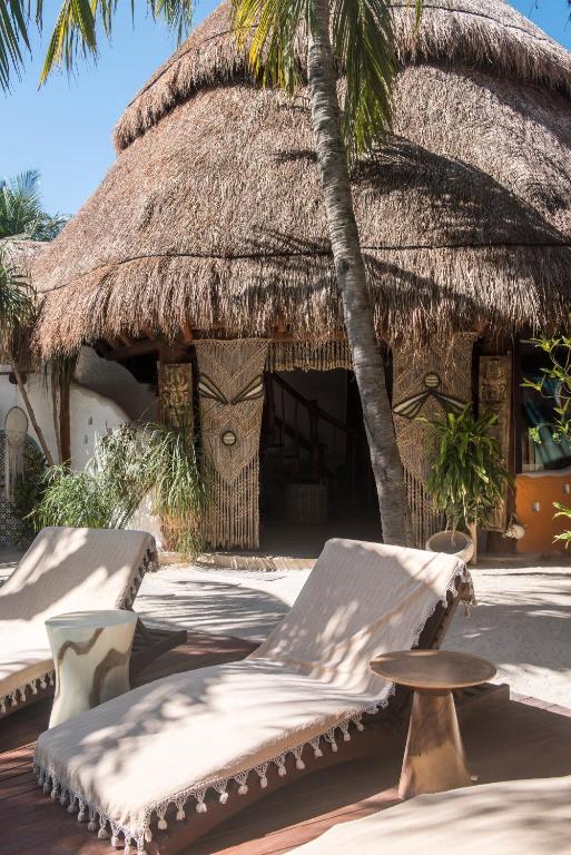 Two beige lounge chairs and a small table sit on a wooden deck under a palm tree in front of a rustic building with a thatched roof and decorative beaded curtains, surrounded by tropical plants.