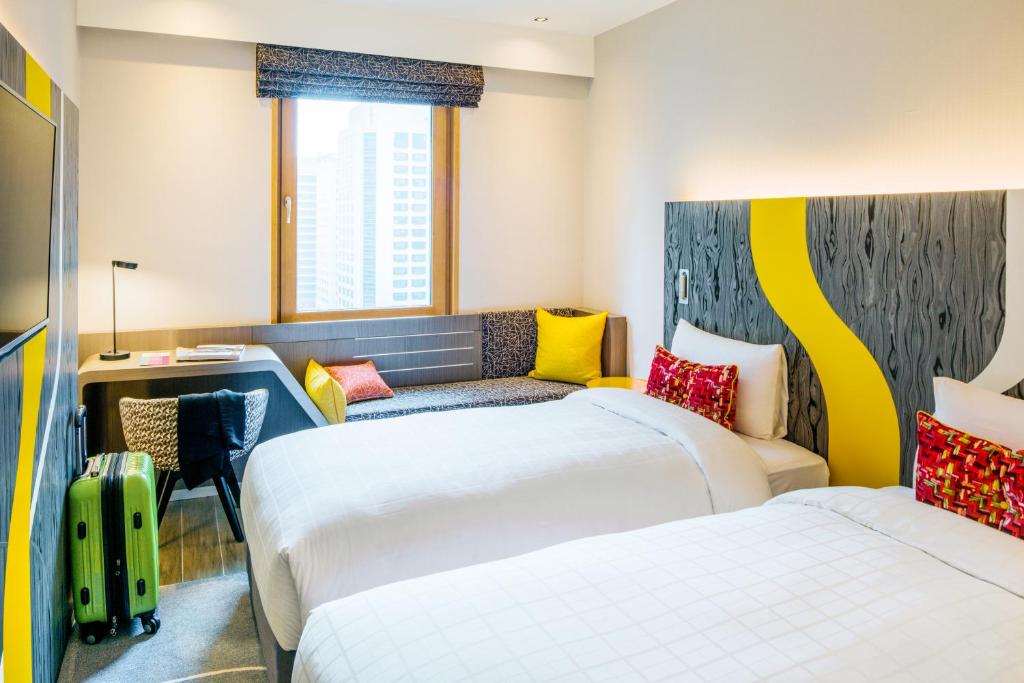Modern hotel room with two twin beds, colorful headboards, a window with a city view, a desk and chair, a green suitcase, and a bench seat with bright pillows by the window.