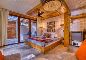 A cozy, modern bedroom with wooden accents, a large bed with gray and red bedding, woven light fixtures, wall art, a rug, and large windows opening to a patio with a small pool.