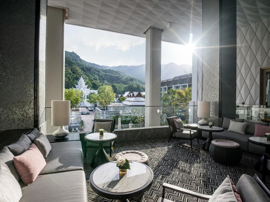 A modern, stylish lounge with sofas and round tables overlooks a scenic view of lush green mountains, trees, and white temple-like buildings under a bright, sunny sky.