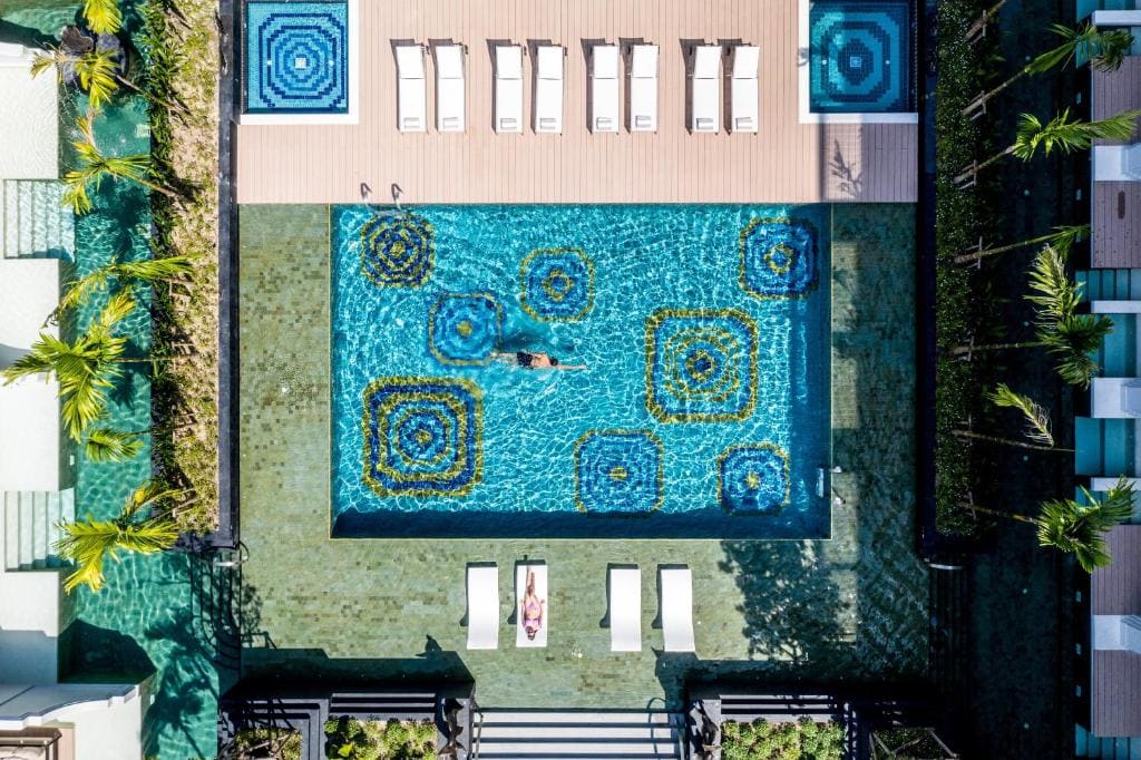 Aerial view of a modern pool with geometric tile patterns. Several white lounge chairs surround the pool, with a person swimming and another sunbathing. Palm trees and greenery line the pool area.