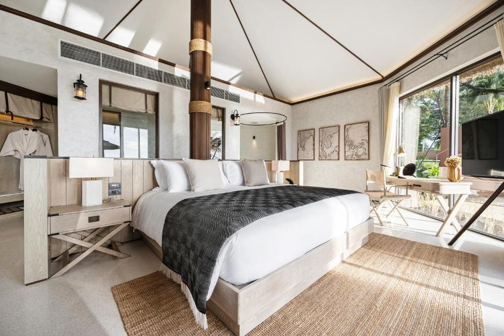 A bright, modern hotel room with a large bed, neutral decor, floor-to-ceiling windows, a small desk and chair, framed artwork on the wall, and natural light streaming in.