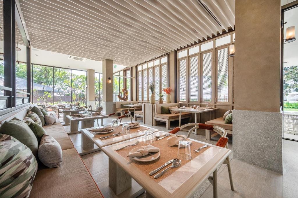A bright, modern restaurant with large windows, wooden tables set for dining, cushioned bench seating, and neutral decor accented with green and orange pillows. Natural light fills the spacious, airy room.