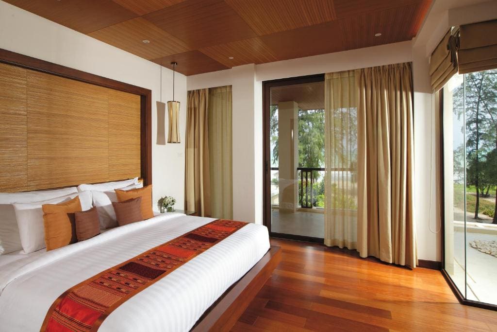 A modern bedroom with wood flooring, a large bed with white linens and orange accent pillows, floor-to-ceiling windows with beige curtains, and a view of trees and a balcony outside.