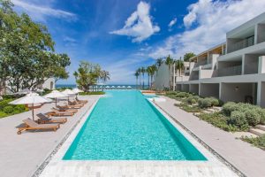 A long, rectangular outdoor pool lined with sun loungers and white umbrellas, surrounded by modern white buildings and lush greenery, leading to a beach and ocean under a partly cloudy sky.