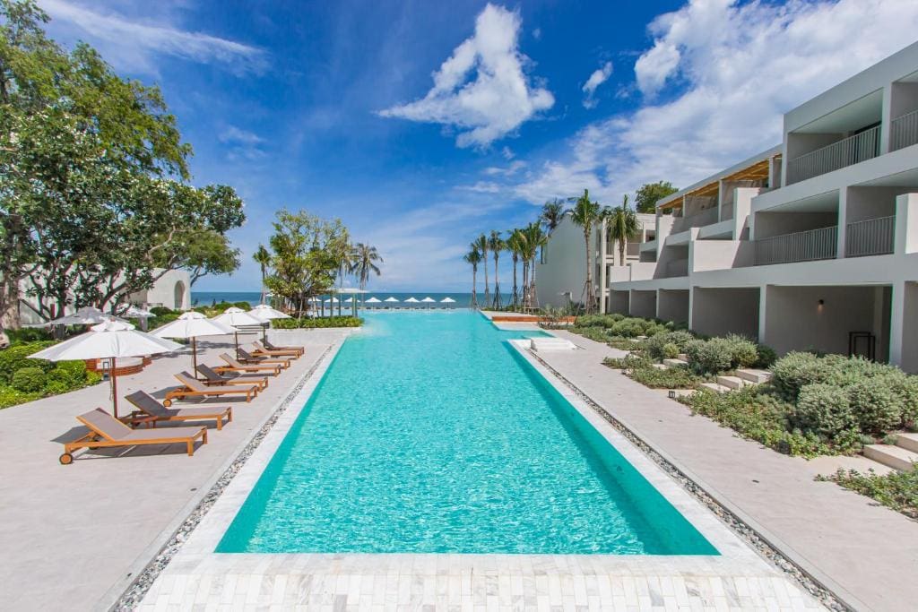 A long, rectangular outdoor pool lined with sun loungers and white umbrellas, surrounded by modern white buildings and lush greenery, leading to a beach and ocean under a partly cloudy sky.
