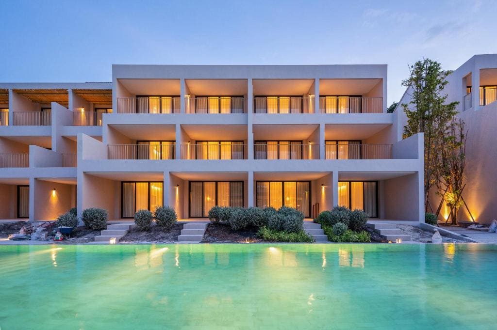 Modern three-story hotel with balconies and large windows, all lit warmly at dusk, overlooks a turquoise swimming pool and is surrounded by greenery and shrubs.