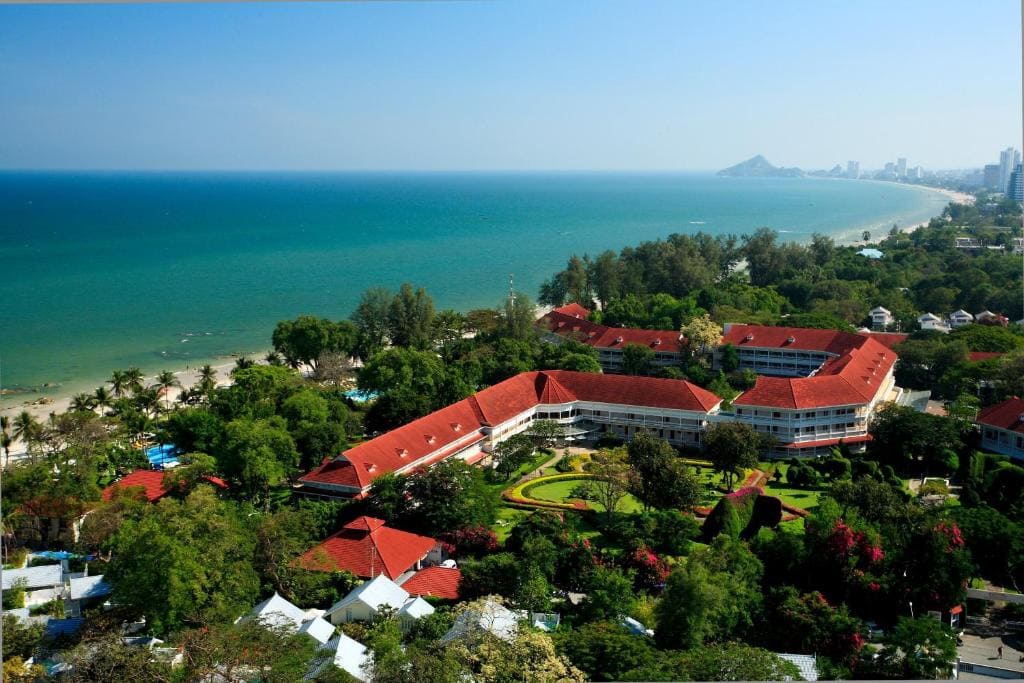 Aerial view of a seaside resort with red-roofed buildings surrounded by lush greenery, overlooking a sandy beach and blue ocean, with a distant city and mountains visible along the coastline.