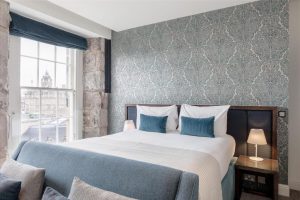 A bright hotel room with a large bed, blue and white bedding, two bedside tables with lamps, patterned wallpaper, and a window showing a cityscape outside.