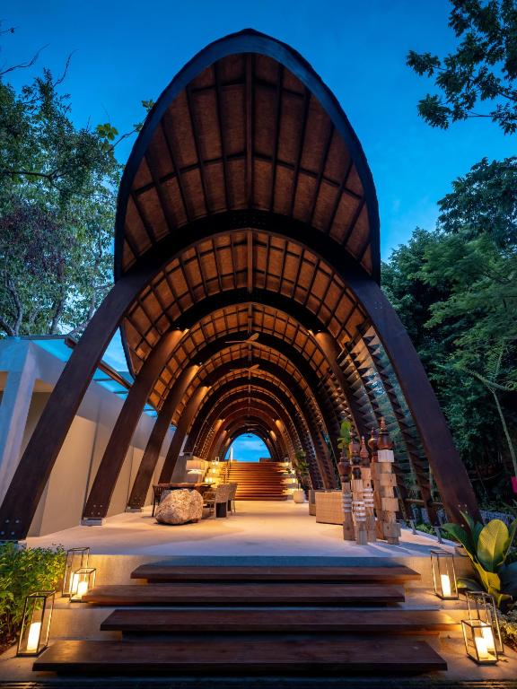 Modern wooden archway entrance with warm lighting, lanterns on steps, and lush greenery on both sides, leading to an open area at dusk with a blue sky visible through the arched roof structure.