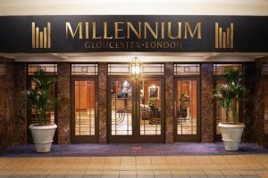 Entrance of Millennium Gloucester Hotel London, featuring glass doors, large potted plants on each side, and a black and gold sign with the hotel’s name above the doors. Warm lighting and an inviting lobby are visible inside.