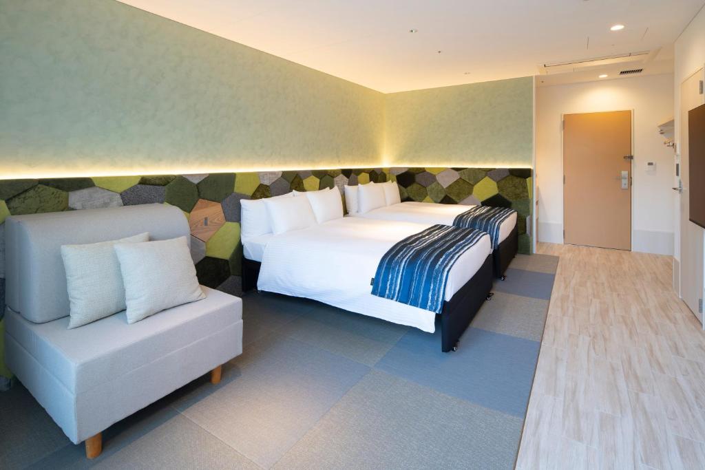 Modern hotel room with two large beds, a geometric green and gray accent wall, a gray cushioned chair with pillows, wood-style flooring, and soft ambient lighting. A wooden door is visible at the far end.