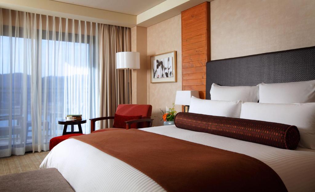 A modern hotel room with a large bed, brown and white bedding, a red armchair, small table, framed art, and floor-to-ceiling window with sheer curtains letting in natural light.