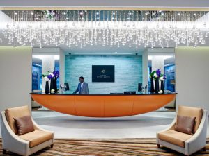 A modern hotel lobby with two cushioned chairs, a curved orange reception desk, and a staff member standing behind it. A decorative chandelier with hanging crystals is above the lobby area.