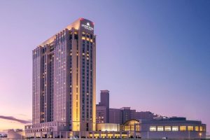 A tall, modern hotel building with “Grand Hyatt” signage illuminated near the top, stands against a clear purple and blue evening sky, with adjacent lower buildings and soft exterior lighting.