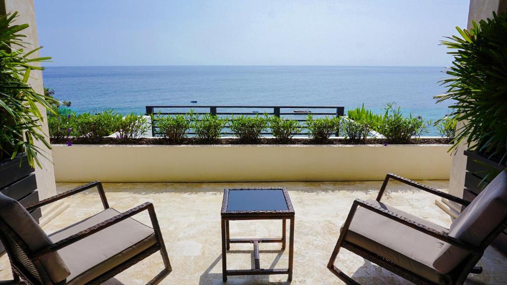 Two cushioned chairs and a small glass-top table sit on a stone patio, overlooking a railing with potted plants and a calm blue ocean under a clear sky in the background.