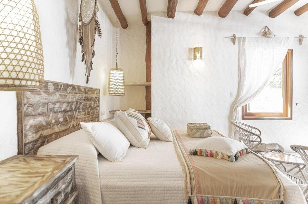 A cozy bedroom with rustic decor, featuring a wooden bed with beige bedding, wicker chairs, woven lamp shades, and a window letting in natural light. Exposed wooden beams and white textured walls add to the warm atmosphere.