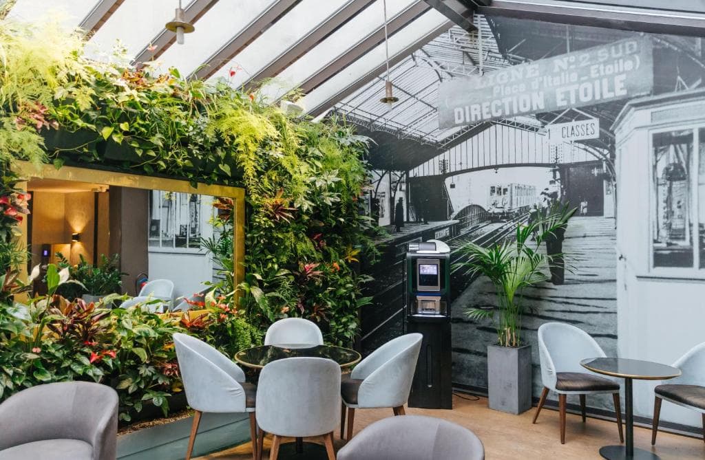 A modern café with gray chairs, glass tables, a large wall mirror surrounded by lush green plants, and a wall mural depicting an old train station with a sign that reads Direction Étoile.