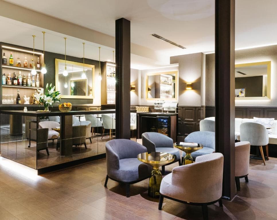 Modern lounge bar with soft armchairs around small round tables, wooden flooring, mirrored walls, shelves of bottles and glasses, decorative lighting, and elegant neutral-toned decor.