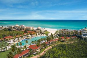 Aerial view of a beachfront resort with large pools, palm trees, and elegant buildings overlooking turquoise ocean water and a white sandy beach under a clear blue sky.