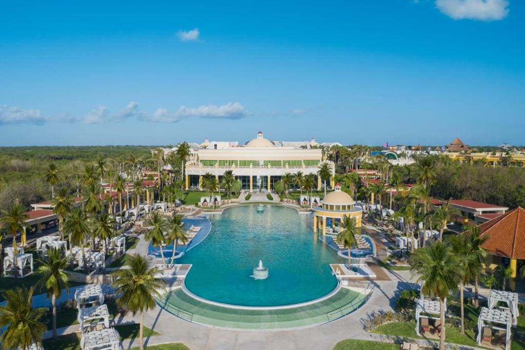 Aerial view of a luxurious resort featuring a large central pool with a fountain, surrounded by palm trees, cabanas, lounge chairs, and elegant buildings under a clear blue sky.
