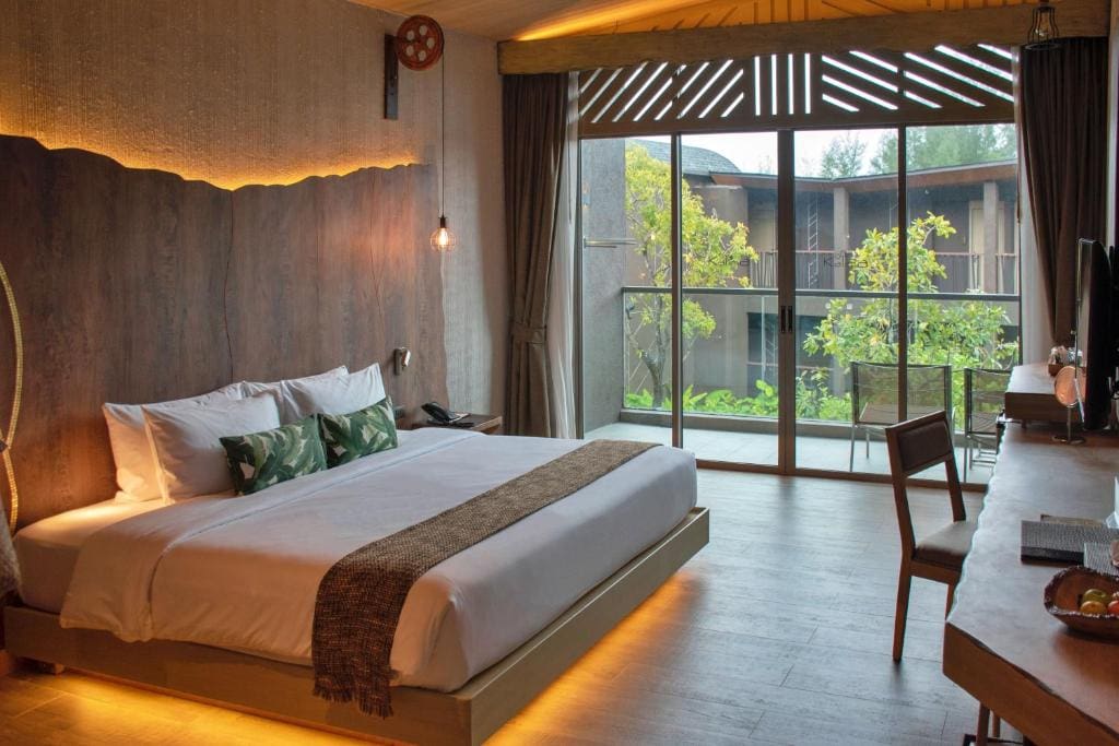 Modern hotel room with a large bed, soft lighting, wooden accents, and a desk. Floor-to-ceiling windows open to a balcony with chairs, overlooking greenery and another building.