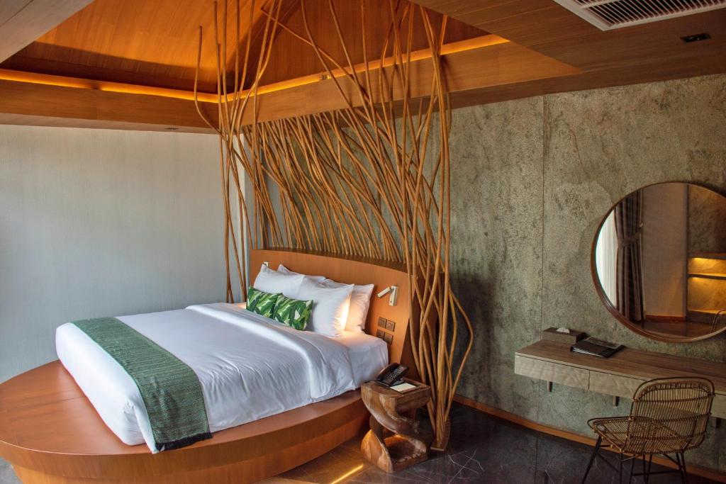 A modern bedroom with a wooden bed frame, white linens, green accents, and decorative wooden branches as a headboard. The room features a round mirror, a wooden desk with a wicker chair, and earthy-toned walls and ceiling.
