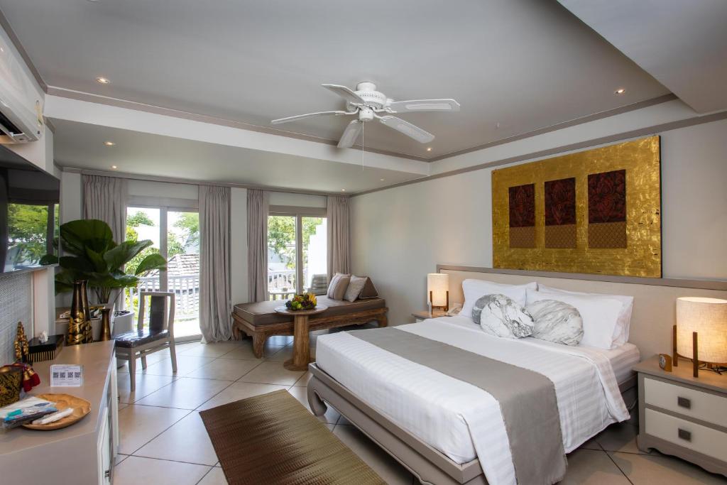 A modern hotel room with a large bed, neutral decor, a ceiling fan, a sitting area with a wooden table, a desk near a window, and gold artwork above the bed. Natural light enters through glass doors to a balcony.