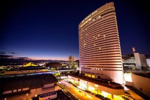A tall, modern hotel building with curved architecture is brightly lit against a deep blue evening sky, overlooking a cityscape with lights and several other buildings.