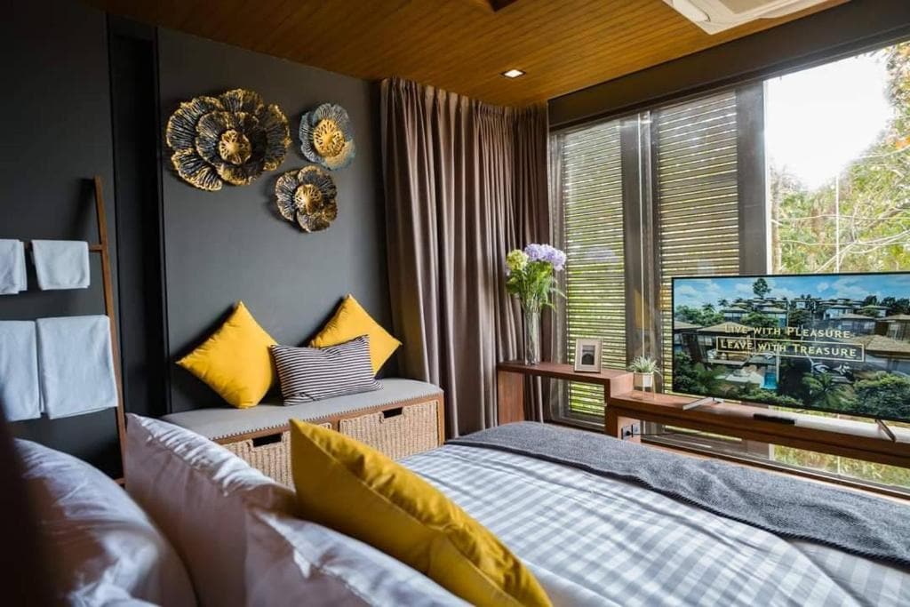 A cozy modern bedroom with a bed covered in plaid sheets and yellow pillows, wall art, a small bench with cushions, large windows with curtains, a vase of flowers, and a TV displaying a scenic image.