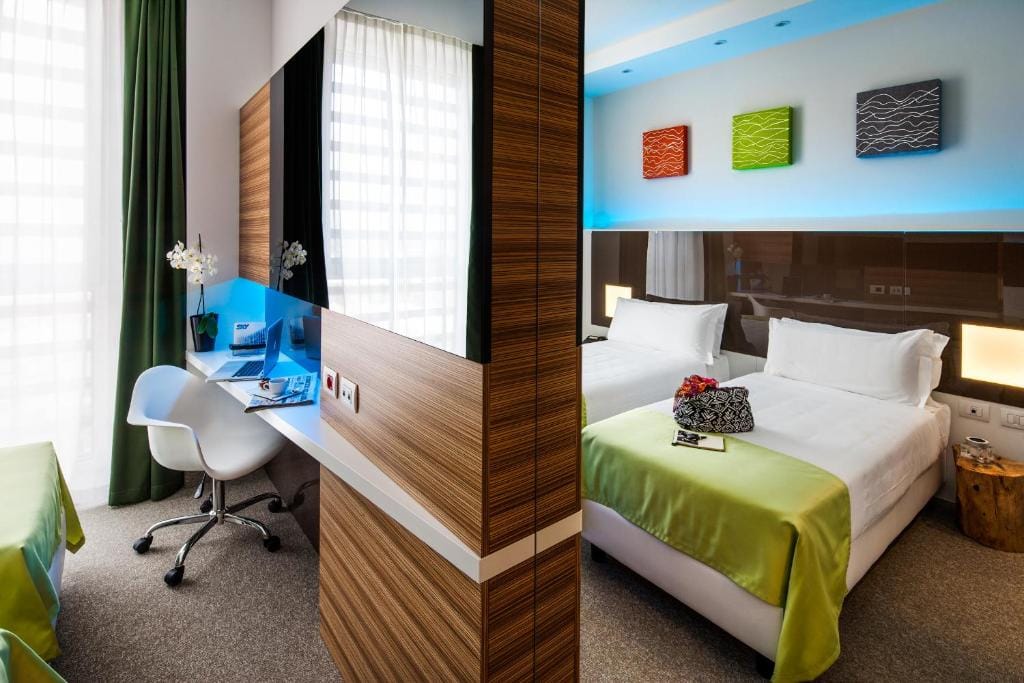Modern hotel room with two beds covered in white linens and green throws, a desk area with a white chair, and colorful wall art. Natural light comes through sheer curtains, and blue accent lighting brightens the room.