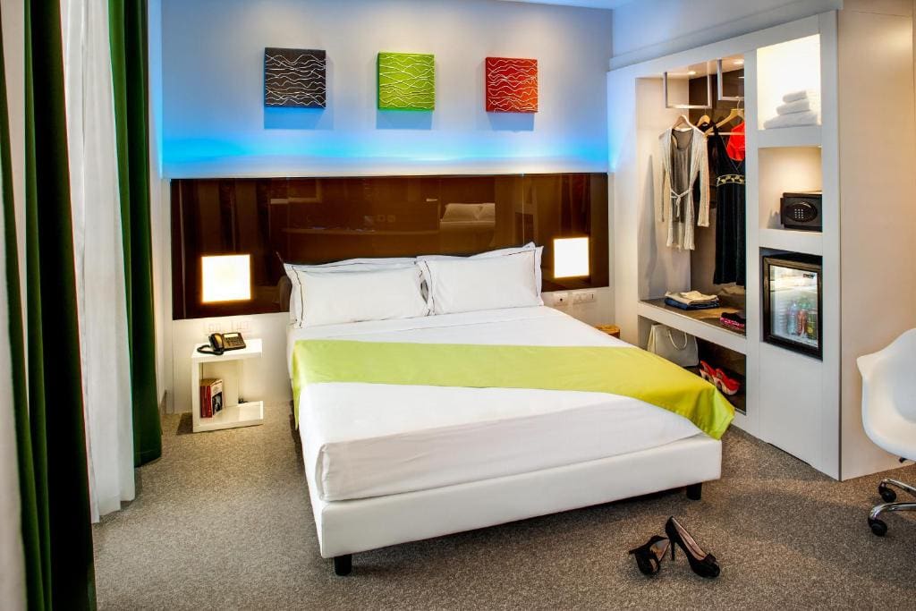 Modern hotel room with a double bed, green and white bedding, open closet, mini fridge, clothes, and shoes on the floor. Three colorful abstract artworks hang above the bed, and the room is brightly lit.