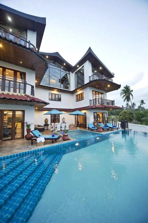 A luxurious modern villa with multiple balconies and large glass windows, featuring an infinity pool with blue tiles, sun loungers, and palm trees in the background at sunset.