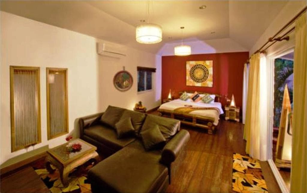 A cozy, warmly lit bedroom with a dark sectional sofa, wooden floors, a large bed against a red accent wall with decorative art, bedside tables, and large windows with sheer curtains overlooking greenery.