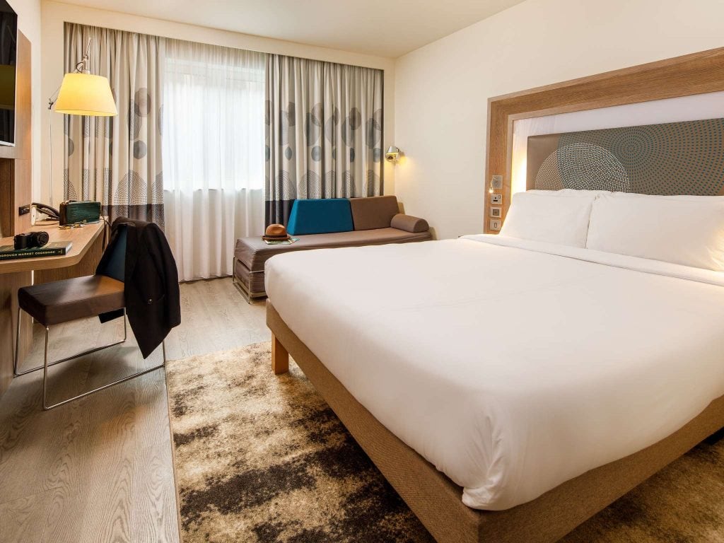 Modern hotel room with a large bed, a desk with a chair, a lamp, a sofa near a window with curtains, and a rug on the floor. The room has neutral tones and simple, contemporary decor.