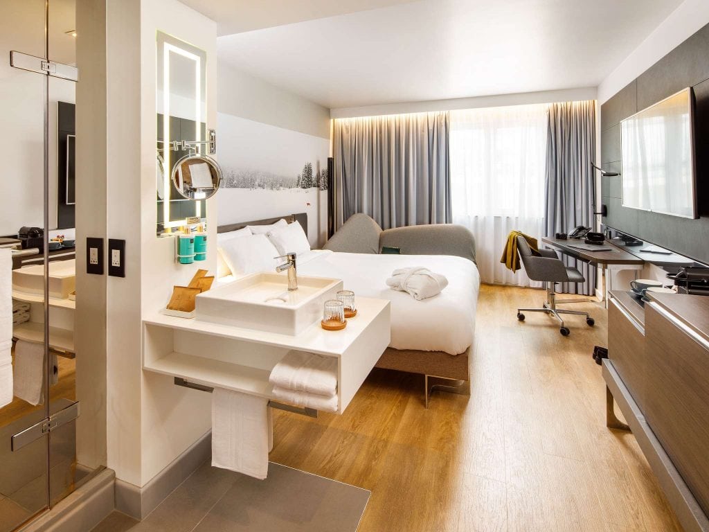 A modern hotel room with a large bed, a minimalist sink area, a flat-screen TV, a desk with a chair, and floor-to-ceiling windows with curtains, featuring wood flooring and contemporary decor.