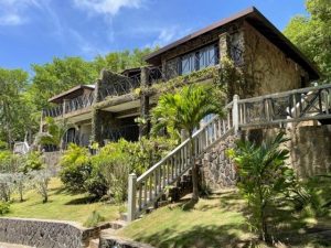 A two-story stone house with balconies, surrounded by lush greenery and palm trees, features a wooden staircase and railing leading up a sloped garden under a bright blue sky.