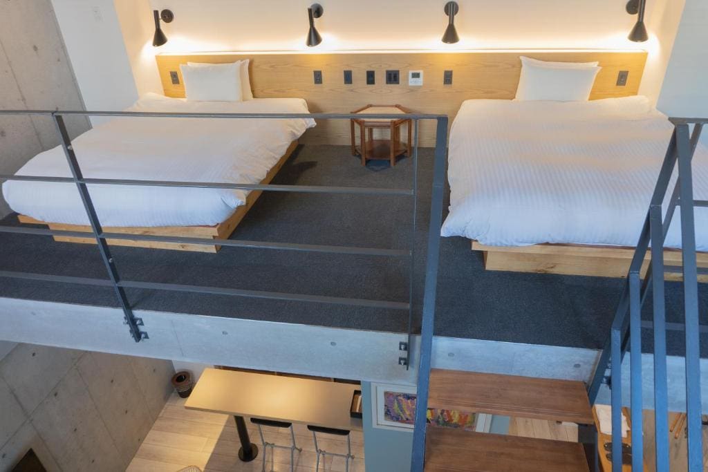 View from a loft showing two beds with white linens on a raised platform, separated by a small wooden table, with black sconces above. Metal railing and stairs lead down to a modern kitchen area below.