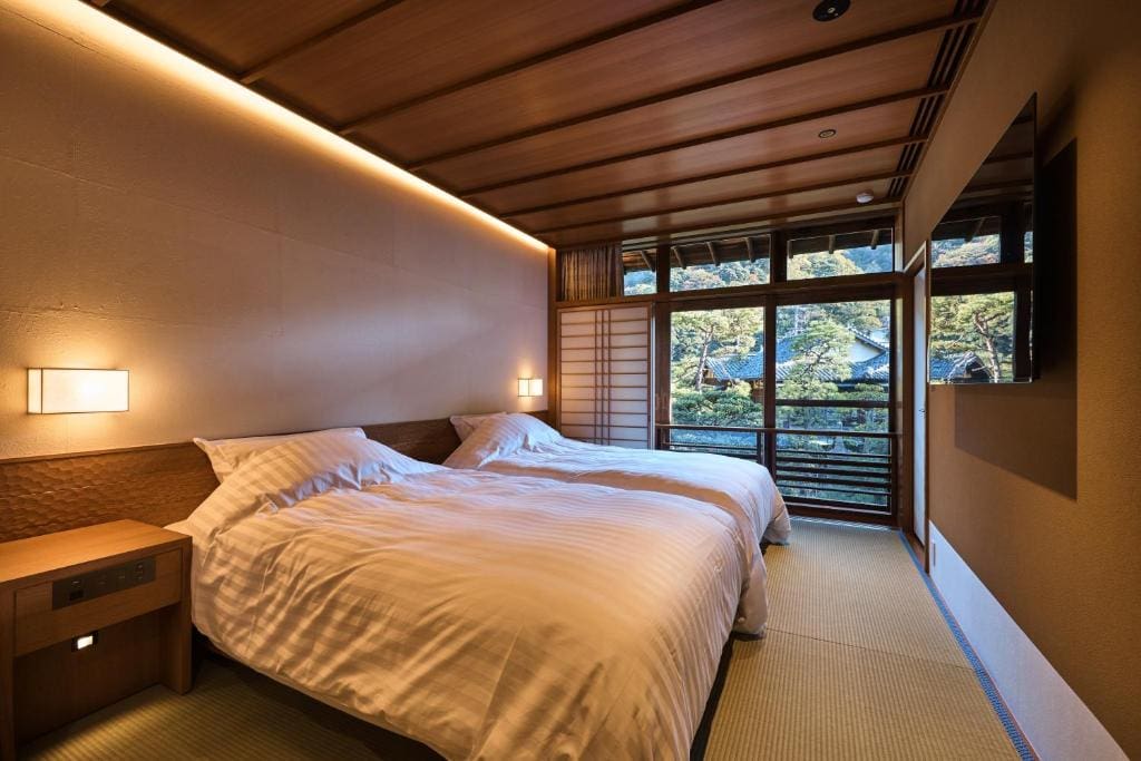 A serene Japanese-style bedroom with two beds, white bedding, tatami mat flooring, wooden ceiling, shoji screens, soft lighting, and a large window overlooking a garden with greenery outside.