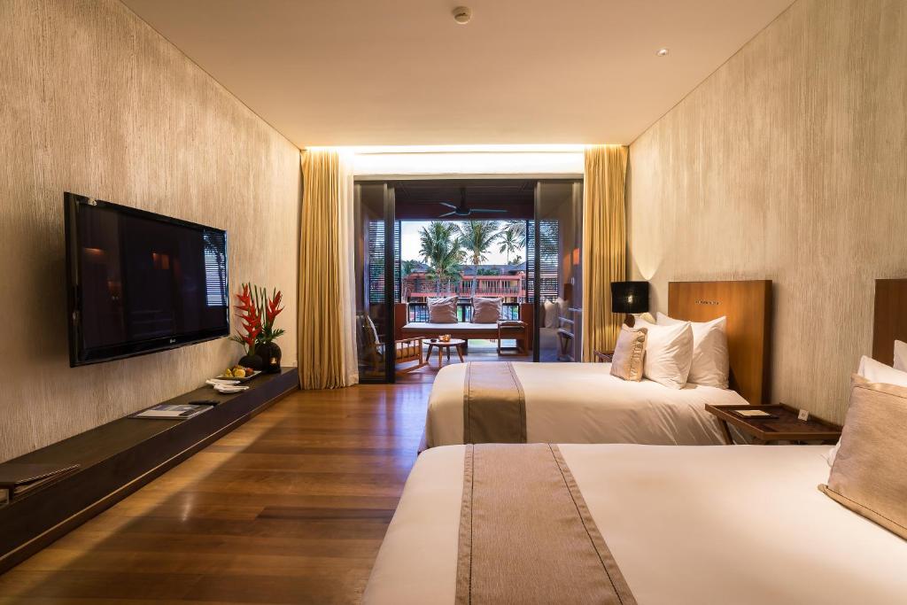 A modern hotel room with two double beds, a flat-screen TV, wooden floors, and large windows with curtains opening to a balcony overlooking palm trees and a pool area outside.