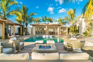 A luxurious outdoor patio with cushioned seating, a glass fire pit, and a swimming pool surrounded by palm trees. There is a poolside bar and lounge area under a shaded roof, all under a bright blue sky.