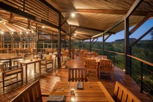 Open-air restaurant with wooden tables and chairs on a polished deck, surrounded by a metal and bamboo roof structure, overlooking greenery and hills at dusk, with warm ambient lighting.