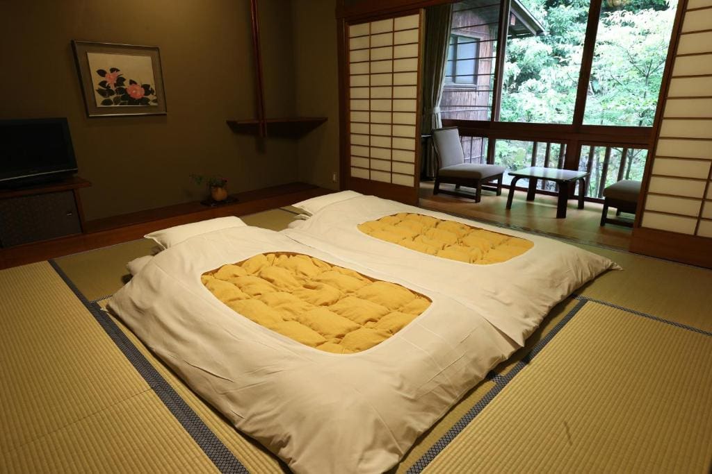 A traditional Japanese tatami room with two futon beds laid on the floor, shoji sliding doors, and a view of a balcony with chairs overlooking a garden.